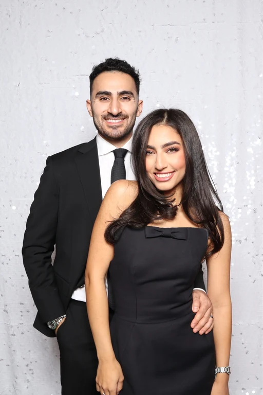 Smiling couple in black formalwear posing against a white sequin backdrop