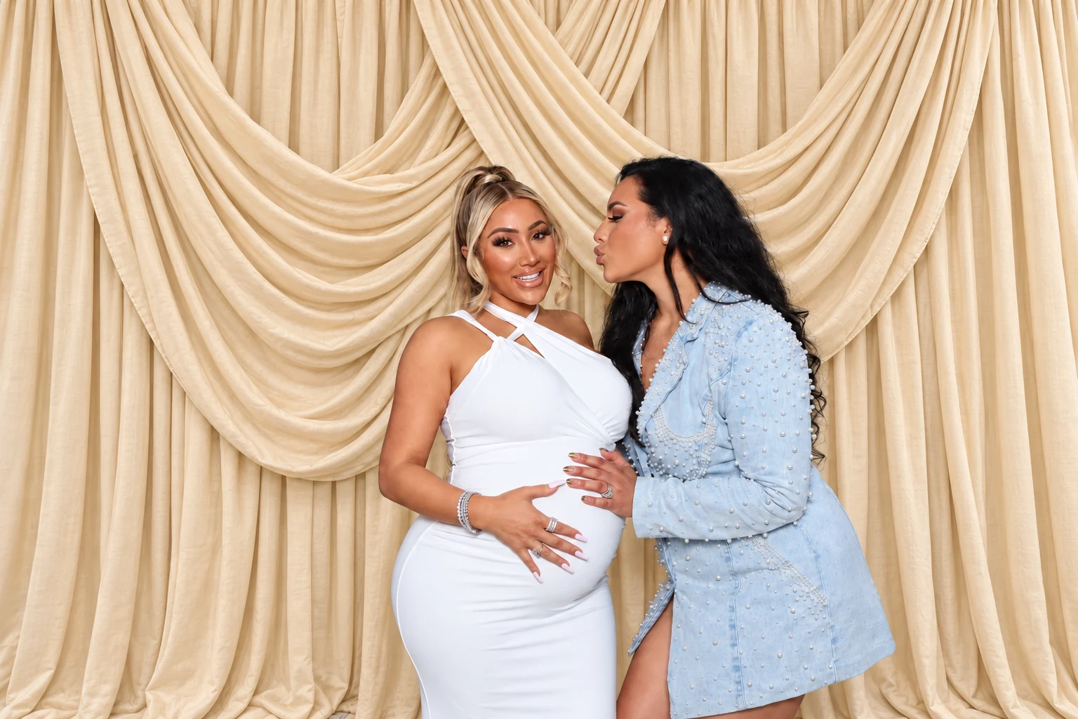 Edited celebration portrait of two women, one in a white dress and one in a light blue outfit, in front of champagne drapes