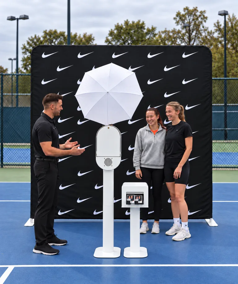 Studio Booth with umbrella flash and printer beside an attendant and two guests in front of a black swoosh-pattern physical backdrop on a tennis court