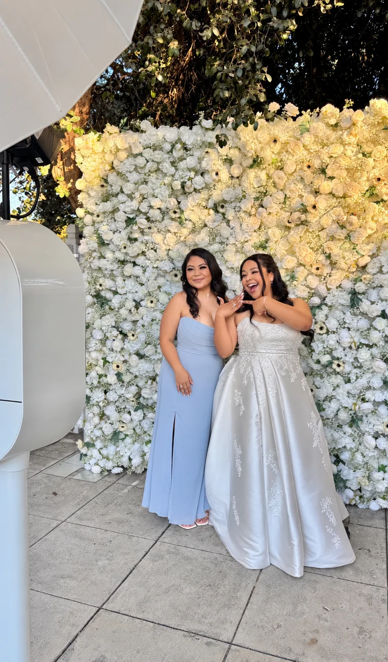 Bride and a bridesmaid in a blue gown posing in front of a white flower wall beside the Studio Booth