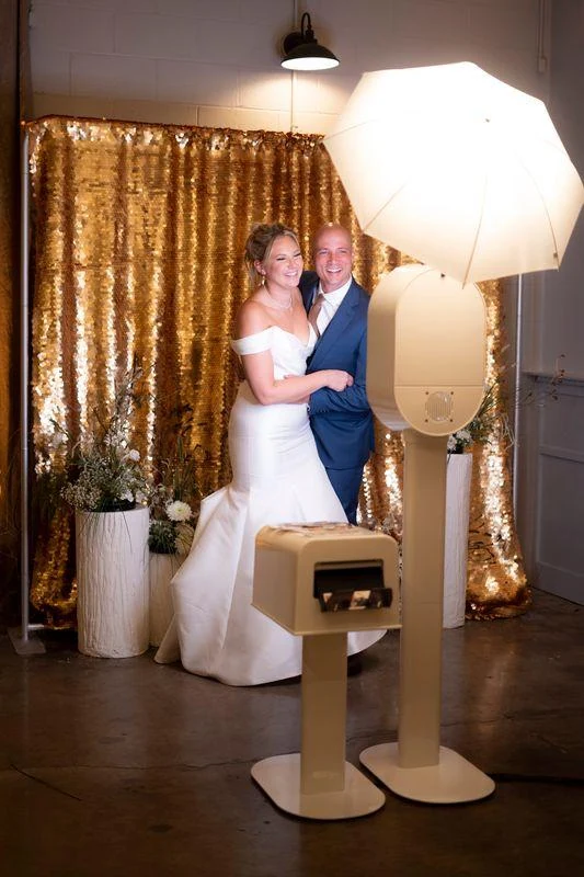 Couple beside a white open-air booth and separate printer in front of a gold sequin backdrop
