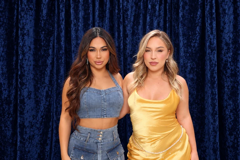Two women in denim and yellow posing together against navy crushed velvet drapes